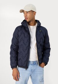 Giacca puffer blu navy con motivo a chevron, cappuccio, chiusura con zip e polsini elastici. Abbinata a jeans chiari strappati e un cappellino beige.