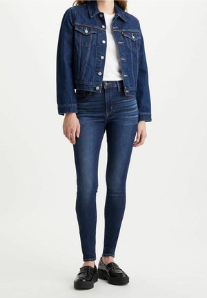 Nainen, jolla on yllään tummansininen farkkutakki, valkoinen t-paita, tummansiniset skinny-farkut ja mustat korolliset loafersit, seisoo valkoista taustaa vasten.