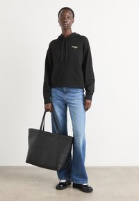 Haut à capuche noir court avec des cordons, associé à des jeans évasés bleu clair. Un sac fourre-tout noir avec deux poignées, sur un fond neutre.