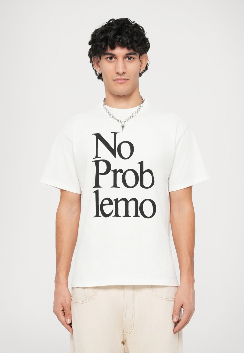 T-shirt bianca in cotone con maniche corte, con la scritta nera in bold "No Problemo" centrata sul davanti. Abbinata a pantaloni beige chiaro.