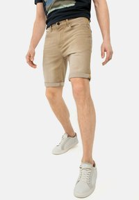 Shorts en denim beige avec des ourlets repliés, des poches avant et une étiquette logo discrète. Associés à des baskets blanches et une chemise à manches courtes sombre.