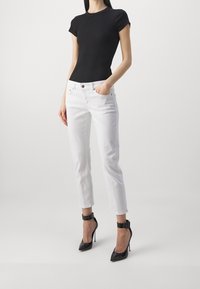 Musta istuva lyhythihainen toppi yhdistettynä valkoisiin suoriin farkkuihin. Mallilla on päällään mustat kiiltävät korkokengät, joissa on nilkkaremmi. Sileät, kiiltävät pinnat.