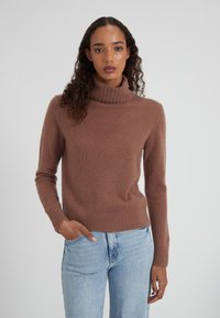Donna che indossa un maglione a collo alto marrone e jeans azzurri chiari, in piedi con una mano in tasca su uno sfondo neutro.