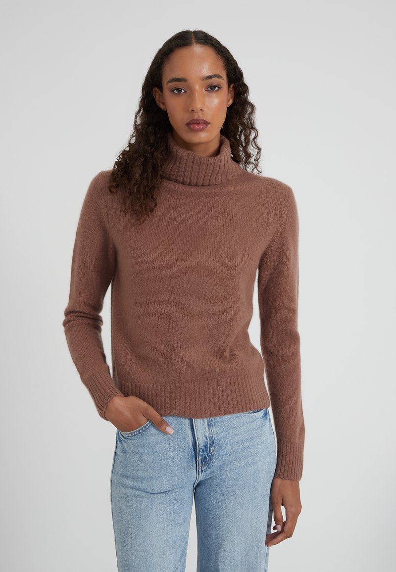 Donna che indossa un maglione a collo alto marrone e jeans azzurri chiari, in piedi con una mano in tasca su uno sfondo neutro.