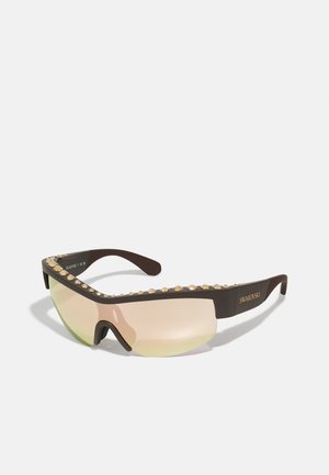 Lunettes de soleil avec une monture marron, ornées de clous dorés sur le dessus, verres dégradés teintés de jaune, design élégant et sportif.