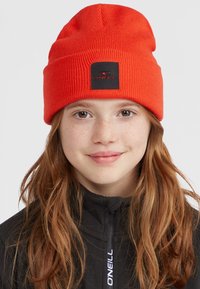 Red ribbed beanie featuring a black square patch with white text. Soft texture. Worn with a zip-up black jacket.