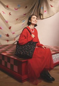 Moteris raudonu megztiniu ir sijonu sėdi ant raudonos languotos staltiesės stalo, laikydama juodą krepšį su žvaigždžių raštu ir šypsodamasi žvaigždėtame fone.