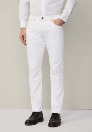 Pantaloni bianchi in cotone con gamba dritta, cinque tasche e chiusura con bottone, abbinati a scarpe in pelle marrone e una camicia chiara.