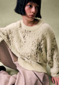 Kobieta o krótkich czarnych włosach, ubrana w kremowy, puszysty sweter z ciemnymi plamkami oraz luźne różowe spodnie, siedząca na tle jednolitego jasnego zielonego tła.