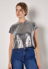 Silberne Pailletten Kurzarmbluse mit runder Halslinie, cropped Design und glatter Textur, kombiniert mit hoch geschnittenen blauen Jeans.