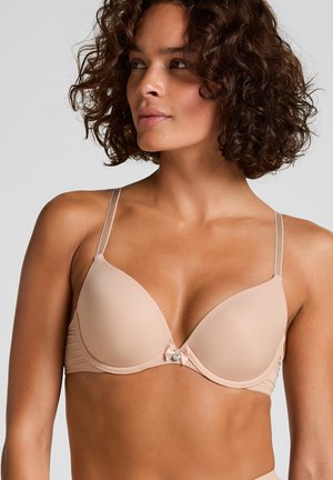 Vrouw met krullend haar die een beige gevoerde beha draagt met dunne bandjes en een klein decoratief strikje op een effen lichte achtergrond.