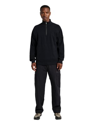 Ung mand stående iført sort halv-zip sweater, sorte cargobukser og beige og sorte sneakers mod en hvid baggrund.