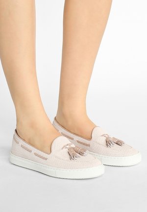 Beige vævede slip-on sko med frynser, båret på bare fødder, med hvide gummisåler og dekorative læderlignende detaljer.