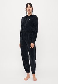 Tuta in velour nero con cappuccio con zip, cintura elastica e polsi elastici. Presenta un piccolo logo sul petto. Texture morbida.