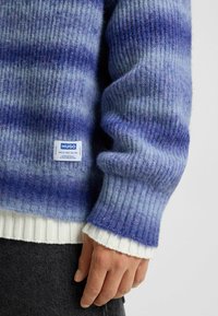 Strikket sweater i blå gradienttoner med ribbet tekstur. Har en hvid nederkant og et mærke. Blødt, hyggeligt materiale.