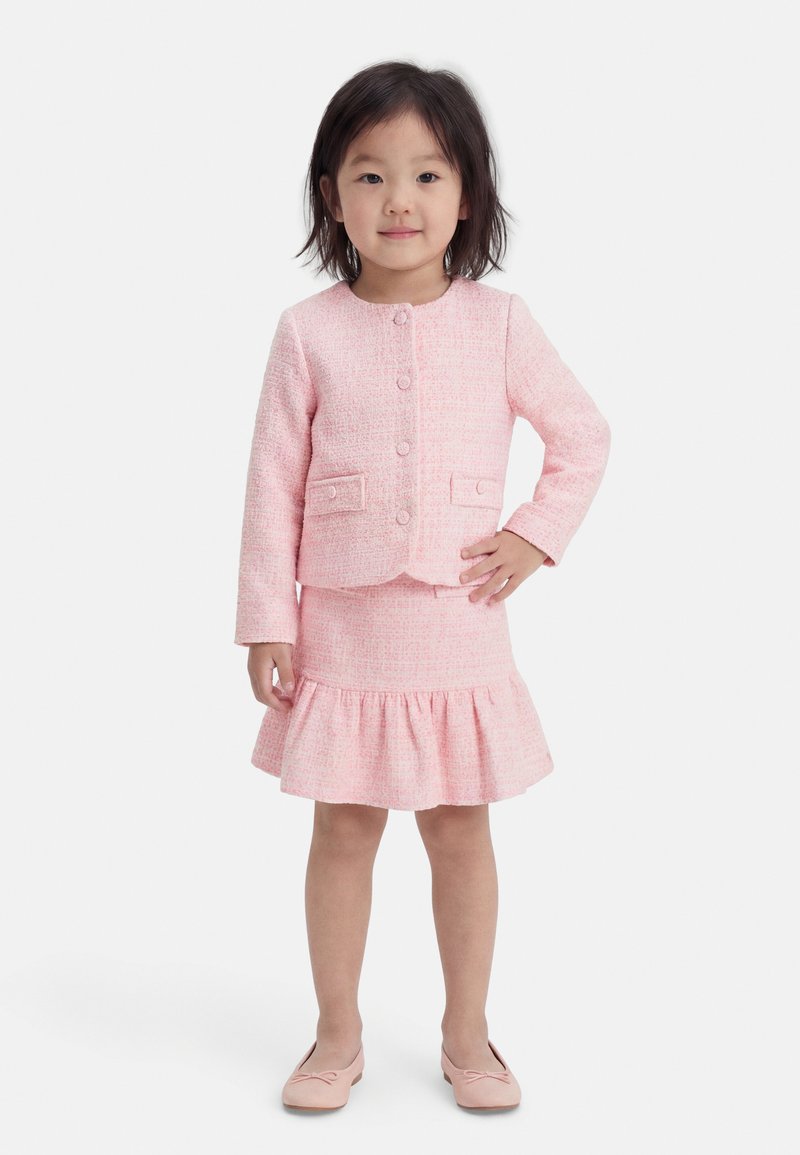 Νεαρή κοπέλα όρθια με το χέρι στη μέση, φοράει ένα ροζ σακάκι με κουμπιά και ταιριαστή φούστα με βολάν και μπαλαρίνες, με φόντο λευκό.