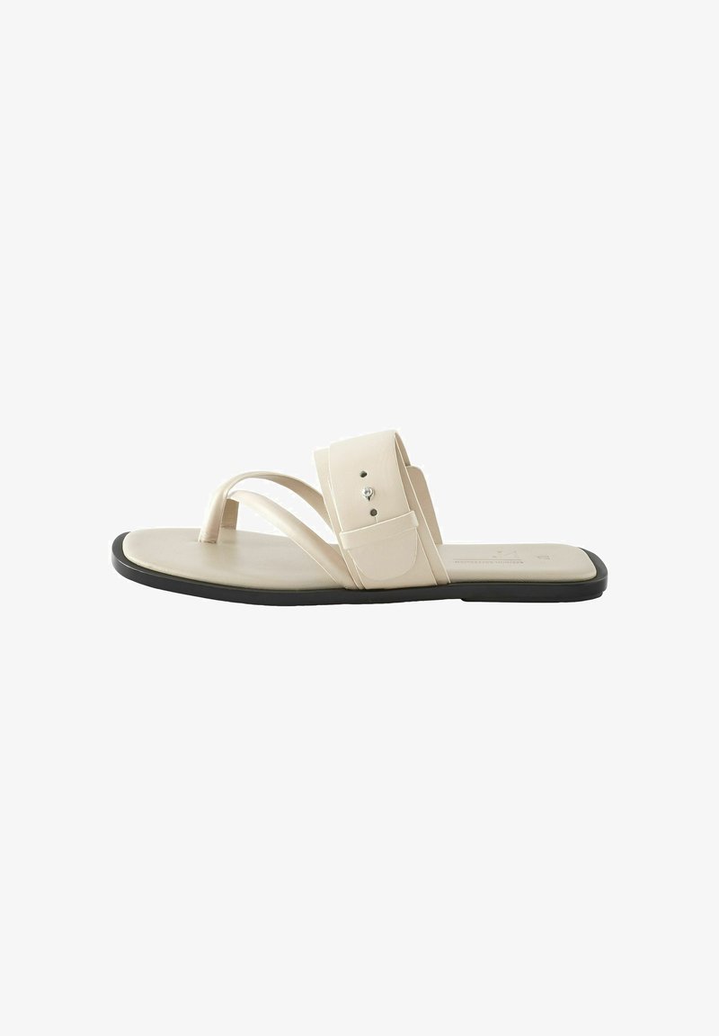 Sandale plate beige avec deux brides croisées et une bride latérale, fabriquée dans un matériau lisse, arborant un design minimaliste et une semelle noire.