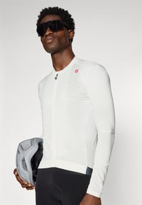 Weißes Langarm-Radtrikot mit strukturierter Oberfläche, durchgehendem Reißverschluss und rotem Logo. Schwarze Radsportshorts und ein grauer Helm sind enthalten.
