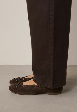 Rudi zomšiniai baleto bateliai su banguotu kraštu ir kaspinėlio detalėmis, dėvimi su tamsiai rudomis tiesiomis kelnėmis ant lygaus grindinio.