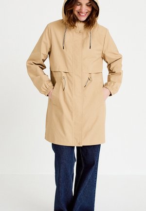 Kobieta w beżowym, ocieplanym płaszczu przeciwdeszczowym z kieszeniami na zamek i niebieskich dżinsach, uśmiechająca się z rękoma w kieszeniach płaszcza na jednolitym tle.