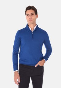 Pullover blu con zip in un design aderente, realizzato in un tessuto morbido con polsini e orlo a coste. Indossato sopra una camicia bianca con colletto.