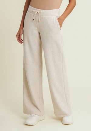 Vrouw die crèmekleurige, wijde joggingbroek met aantrekkoord in de taille en witte sneakers draagt, staand tegen een effen achtergrond.