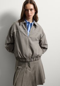 Donna con una giacca grigia con zip e piping sopra una camicia blu con bottoni e una gonna grigia plissettata, in piedi contro una parete bianca semplice.