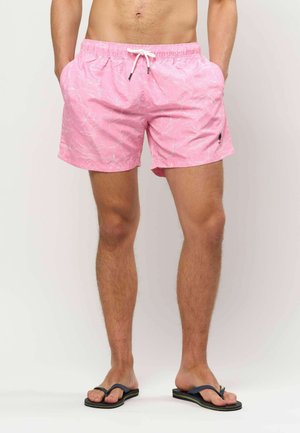 Man draagt roze zwemshorts met bloemenprint, witte trekkoord en zwarte slippers, staat met handen in de zakken tegen witte achtergrond.