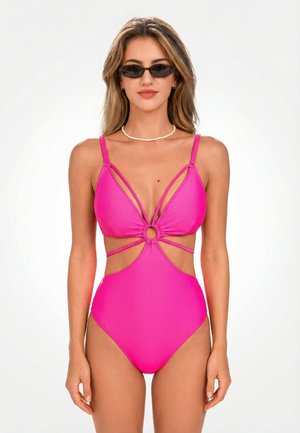 Kobieta ubrana w jasnoróżowy jednoczęściowy strój kąpielowy z wycięciami, czarne wąskie okulary przeciwsłoneczne oraz biały naszyjnik z koralików, stojąca na białym tle.