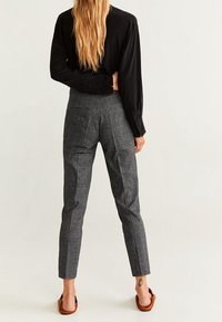 Pantalon à carreaux gris avec une coupe ajustée et des jambes droites, présentant une texture subtile et une taille haute. Porté avec une chemise noire à manches longues.