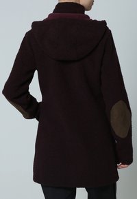 Lång, huva kappa i mörk burgundy med en texturerad yta. Har bruna mockapatchar på armbågarna och en avslappnad passform.