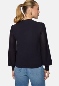 Femme avec une queue de cheval blonde portant un pull bleu marine à manches bouffantes transparentes et un jean bleu clair, de profil sur un fond blanc.
