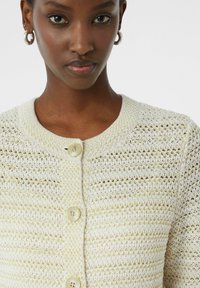 Gebreide cardigan in crème en lichtbeige met een textuurpatroon, voorzien van een ronde hals en vier grote, lichte knopen aan de voorkant.