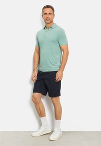 Hellgrünes Poloshirt mit kurzen Ärmeln und zwei Knöpfen, kombiniert mit marineblauen Shorts. Das Modell trägt weiße Sneakers und Socken und steht vor einem schlichten Hintergrund.