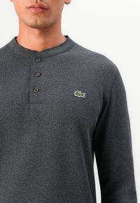 Cămașă henley cu mâneci lungi, de culoare gri închis, cu dungi, prevăzută cu trei nasturi maro și un mic logo verde și alb în formă de crocodil pe partea stângă a pieptului.