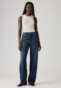 Traperice tamnoplave boje visoka struka s širokim nogavicama, uparene s pripijenim kremastim rebrastim topom bez rukava. Jednostavan dizajn s minimalnim metalnim detaljima.