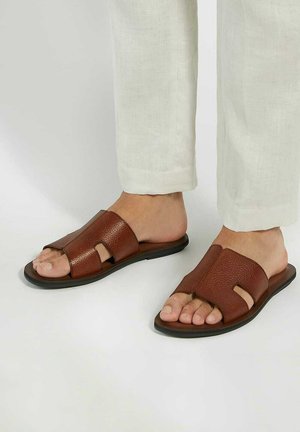 Brown leather slide sandals with an open-toe design, featuring textured straps and cutouts, paired with light-colored linen pants.