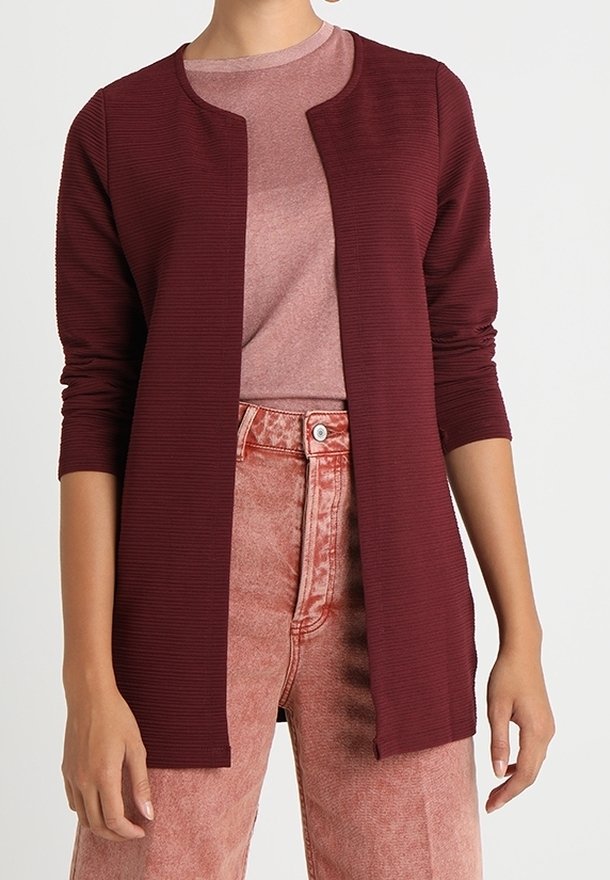 Personne portant un cardigan bordeaux côtelé, ouvert devant, sur un top rose clair et un jean taille haute rose délavé à l’acide, debout devant un fond uni.