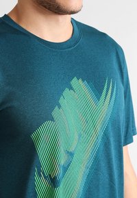 Teal kortärmad t-shirt med rund hals, prydd med ett diagonalgrönt och gult randigt mönster på framsidan. Mjuk, strukturert tyg.