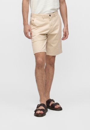 Mand iført beige knælange shorts, hvid skjorte og mørkebrune sandaler med dobbelt rem, stående mod en ensfarvet baggrund.