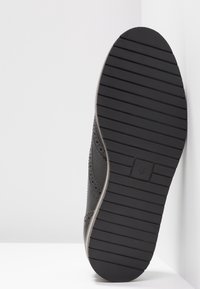 Semelle de chaussure noire avec des rainures horizontales et une légère courbure, reposant contre une surface d'angle blanche.