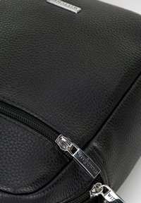Zaino in pelle nera con superficie testurizzata, dotato di doppie cerniere argentate e di un'etichetta del marchio prominente. Design compatto e rettangolare.