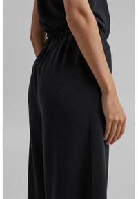 Pantalon noir à jambes larges avec une taille élastique, présentant une texture lisse et une coupe décontractée, vu de profil avec une main posée sur la hanche.