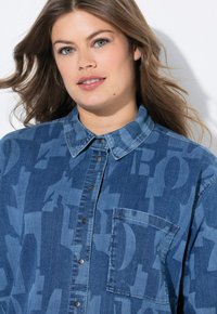 Camicia in denim in diverse tonalità di blu, con abbottonatura frontale, colletto e una singola tasca sul petto con un motivo geometrico.