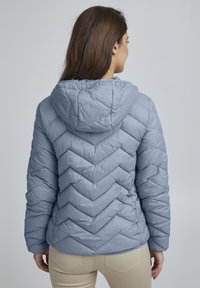 Veste matelassée bleu clair avec un motif en chevrons, une capuche et une texture lisse et brillante. Comprend des manches longues et une silhouette ajustée.