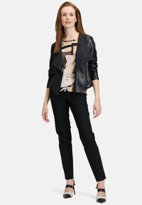 Veste en cuir noir avec un devant drapé sur une blouse imprimée. Le modèle porte un pantalon noir slim et des talons hauts noir et blanc. Texture lisse.