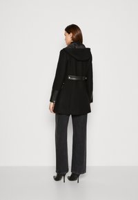 Cappotto nero con cappuccio, caratterizzato da dettagli in pelle sulle maniche e una cintura in vita. Indossato sopra jeans scuri a vita alta e scarpe con tacco alto.