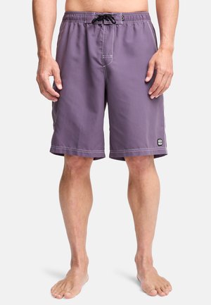 Man draagt paarse zwembroek tot aan de knieën met zwarte koord en een klein logo op het onderste linkerbeen, staat blootsvoets tegen een effen achtergrond.