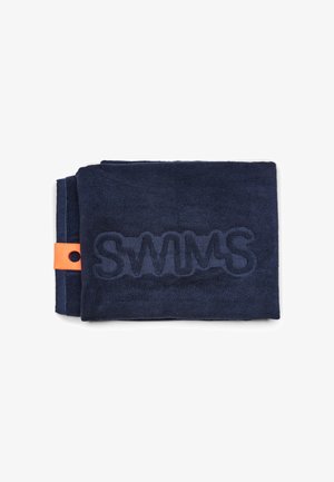 Serviette bleu marine pliée avec « SWIMS » embossé en grandes lettres, étiquette orange sur le côté gauche, fond blanc uni.