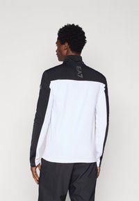 Piklike käised spordijakk valge ja musta värviga, kõrge krae, tekstureeritud kangas ja logo taga. Tavaline lõige kontrastsete varrukatega.